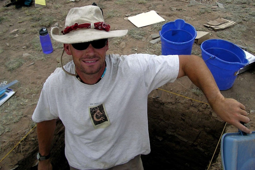 Young scientist in a white hat and shirt is waste deep in a archaeological pit