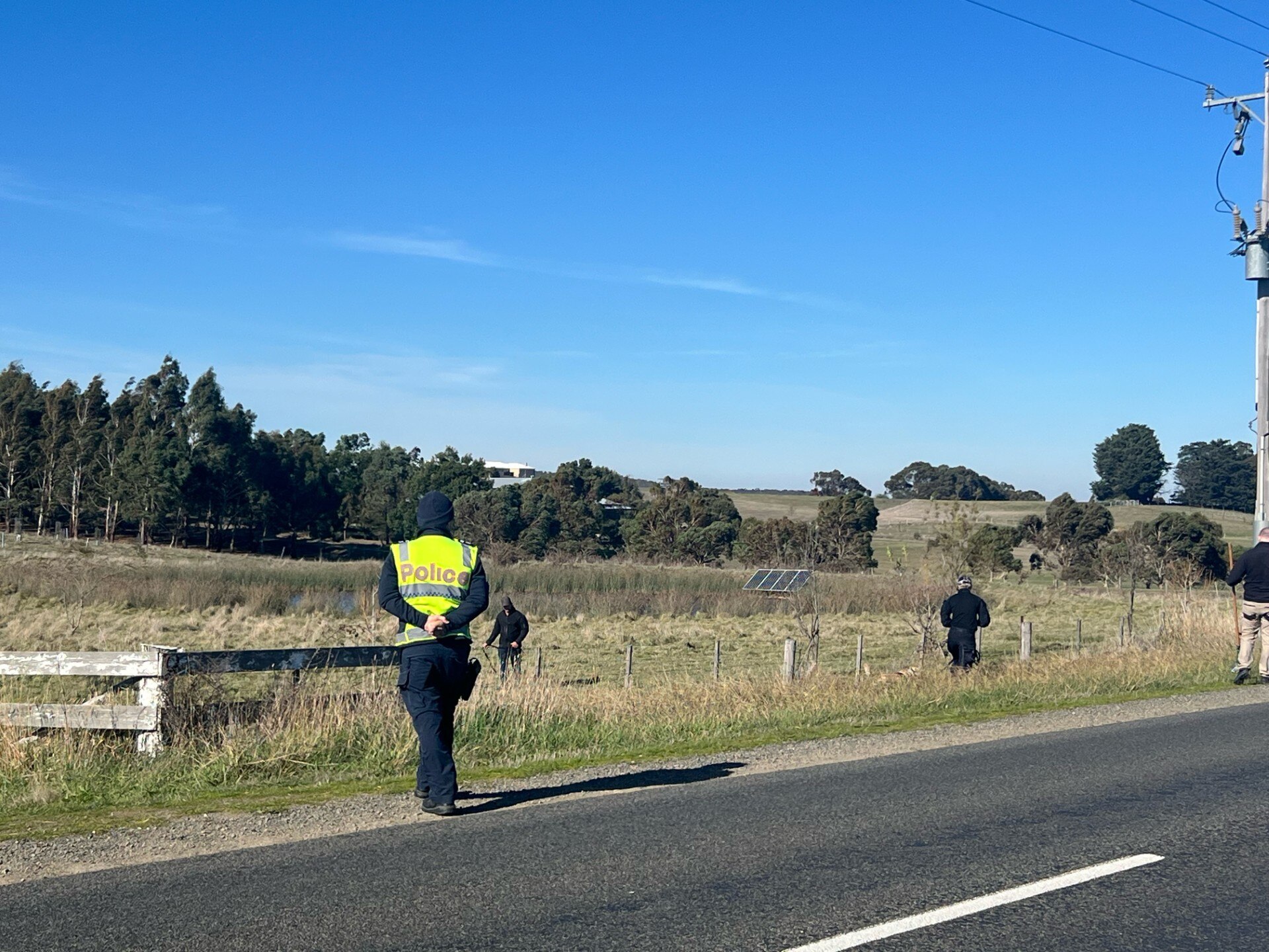 Police officer walking across a road other officers walking through paddocks. 