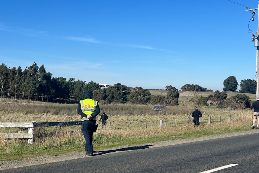 Police officer walking across a road other officers walking through paddocks. 