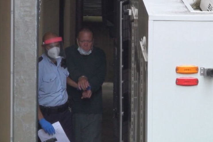 A man in handcuffs is being led by a security guard in a blue shirt and mask.