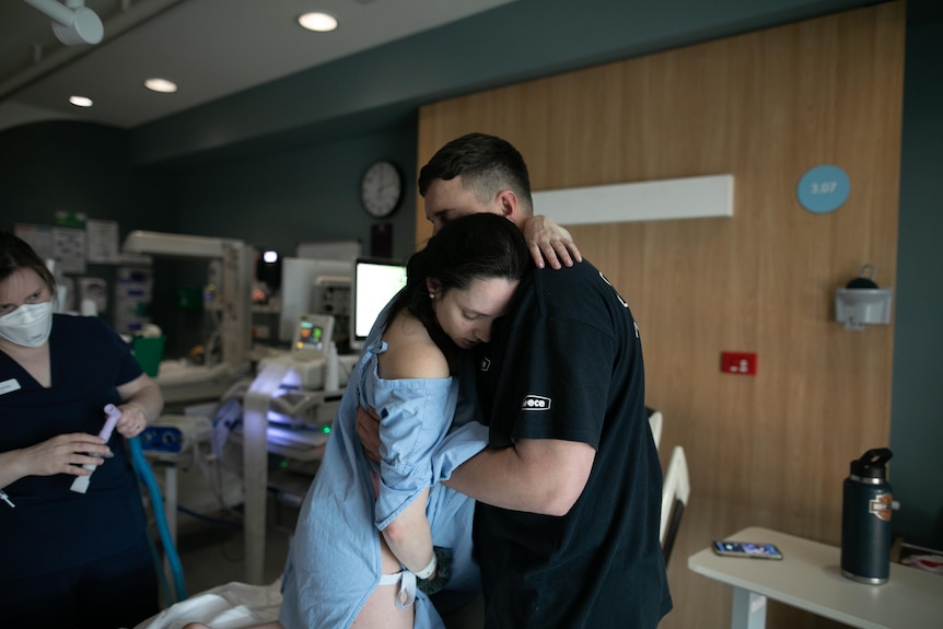 Taryn wears a blue hospital gown, standing up and leaning into the arms of her partner Nathan.