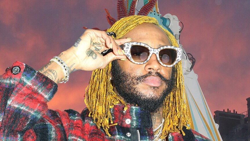 Musician Thundercat wears sunglasses that he holds. He has yellow dreadlocks and a flannel shirt.  