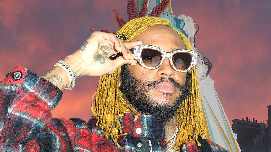 Musician Thundercat wears sunglasses that he holds. He has yellow dreadlocks and a flannel shirt.  