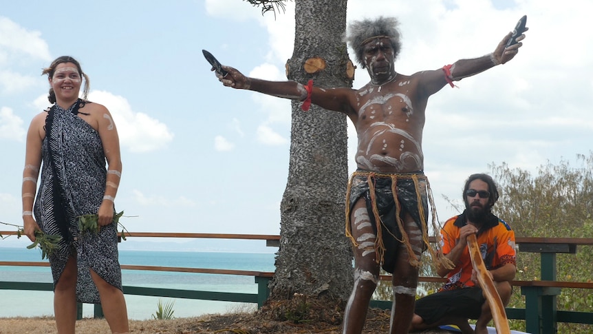 Dancer stands with both arms out holding music sticks and in traditional costume with his body painted