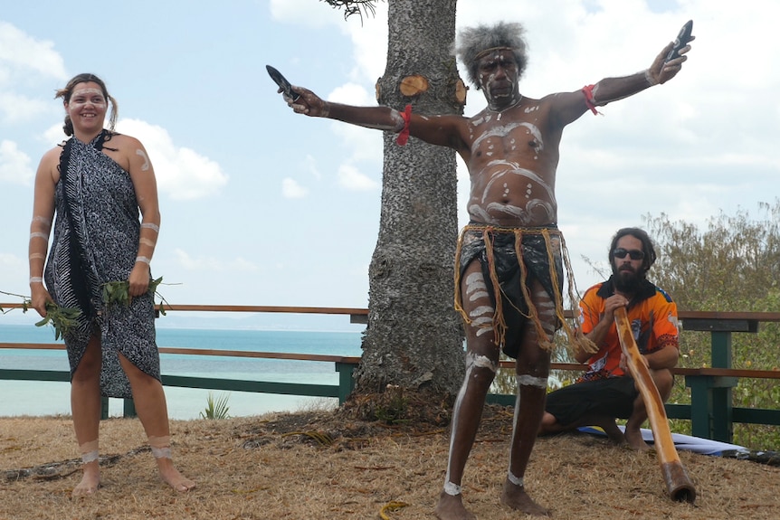 Dancer stands with both arms out holding music sticks and in traditional costume with his body painted