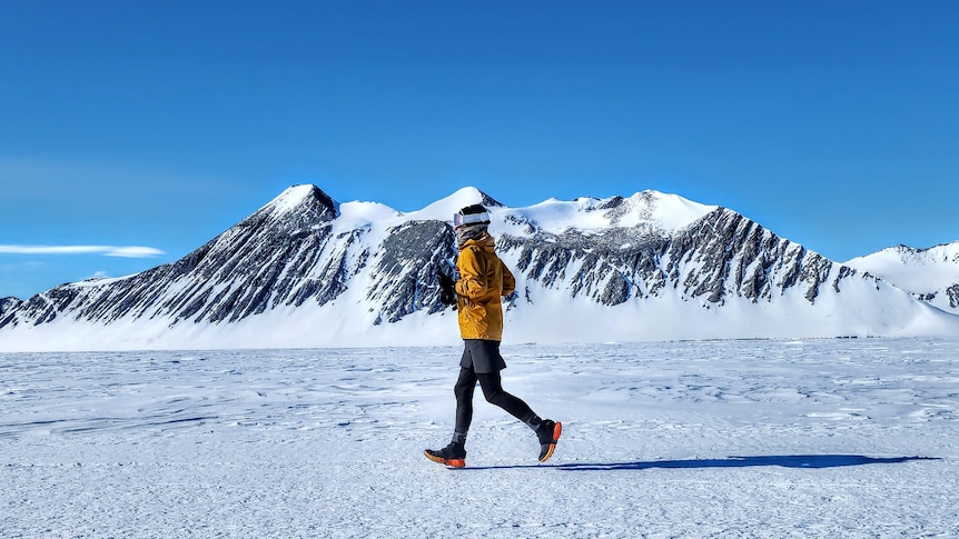 Woman in winter-wear running on snow with ice-capped mountain in background and clear blue sky