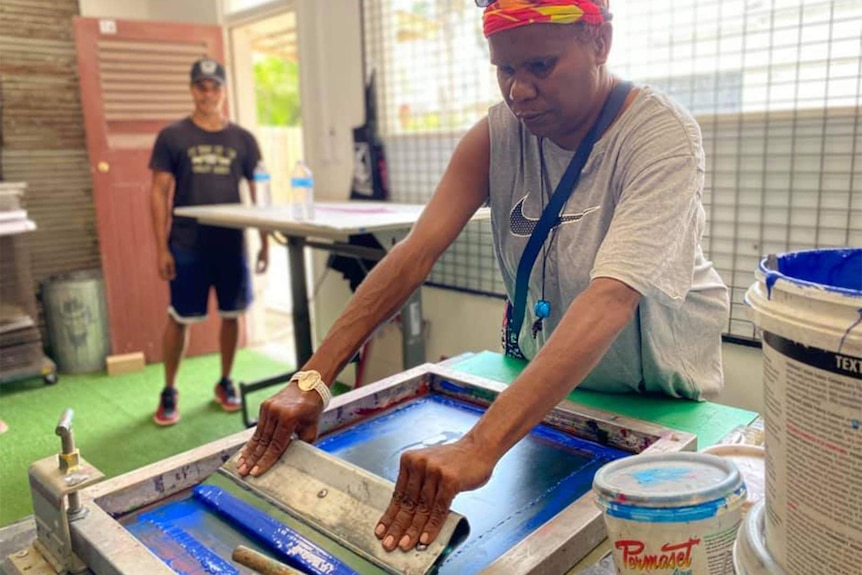 Roxanne Oakley screen printing with blue paint, buckets, table and art supplies behind.