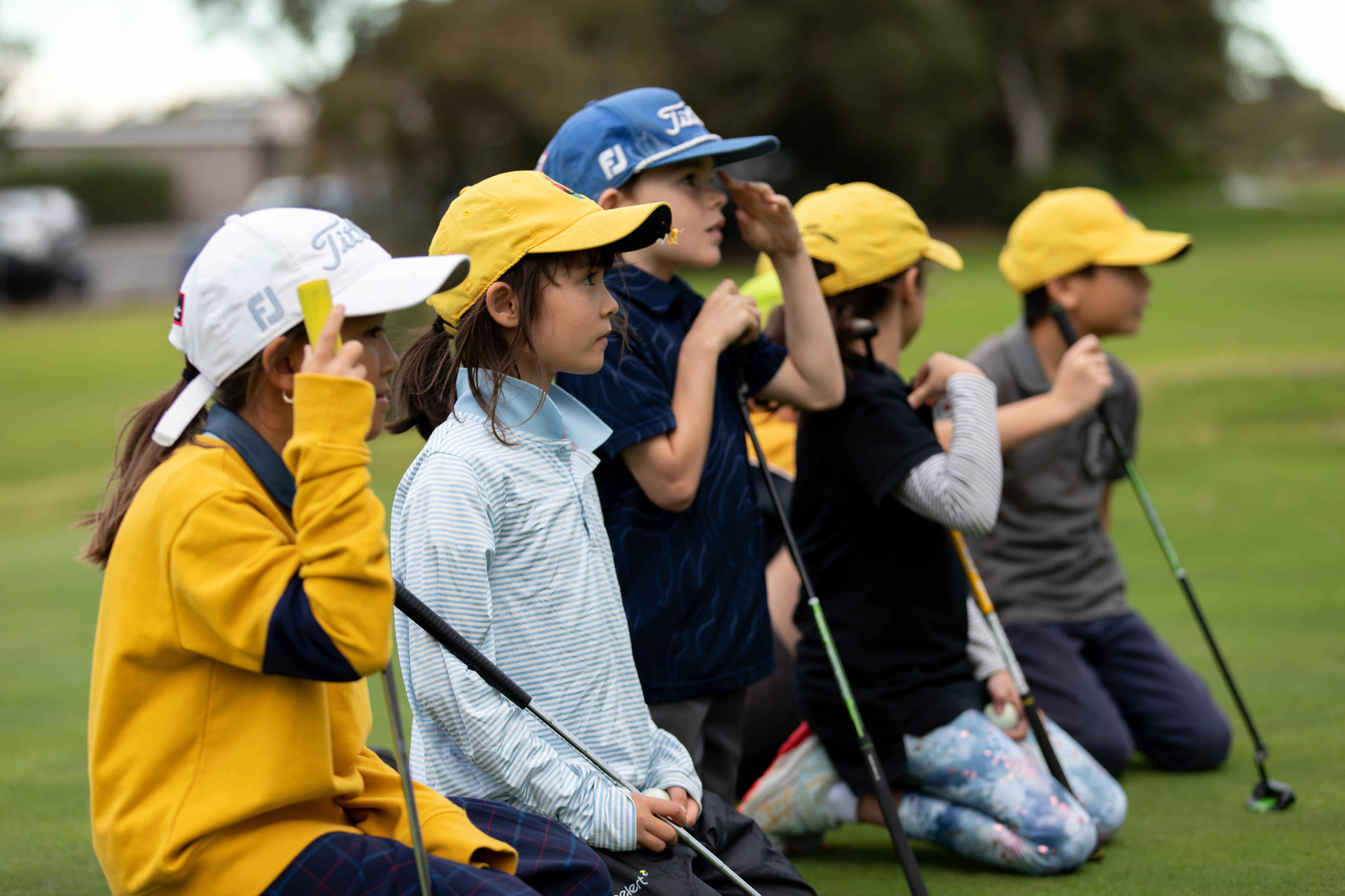 Six children with golf clubs and caps on crouched in a line on a manicured lawn.