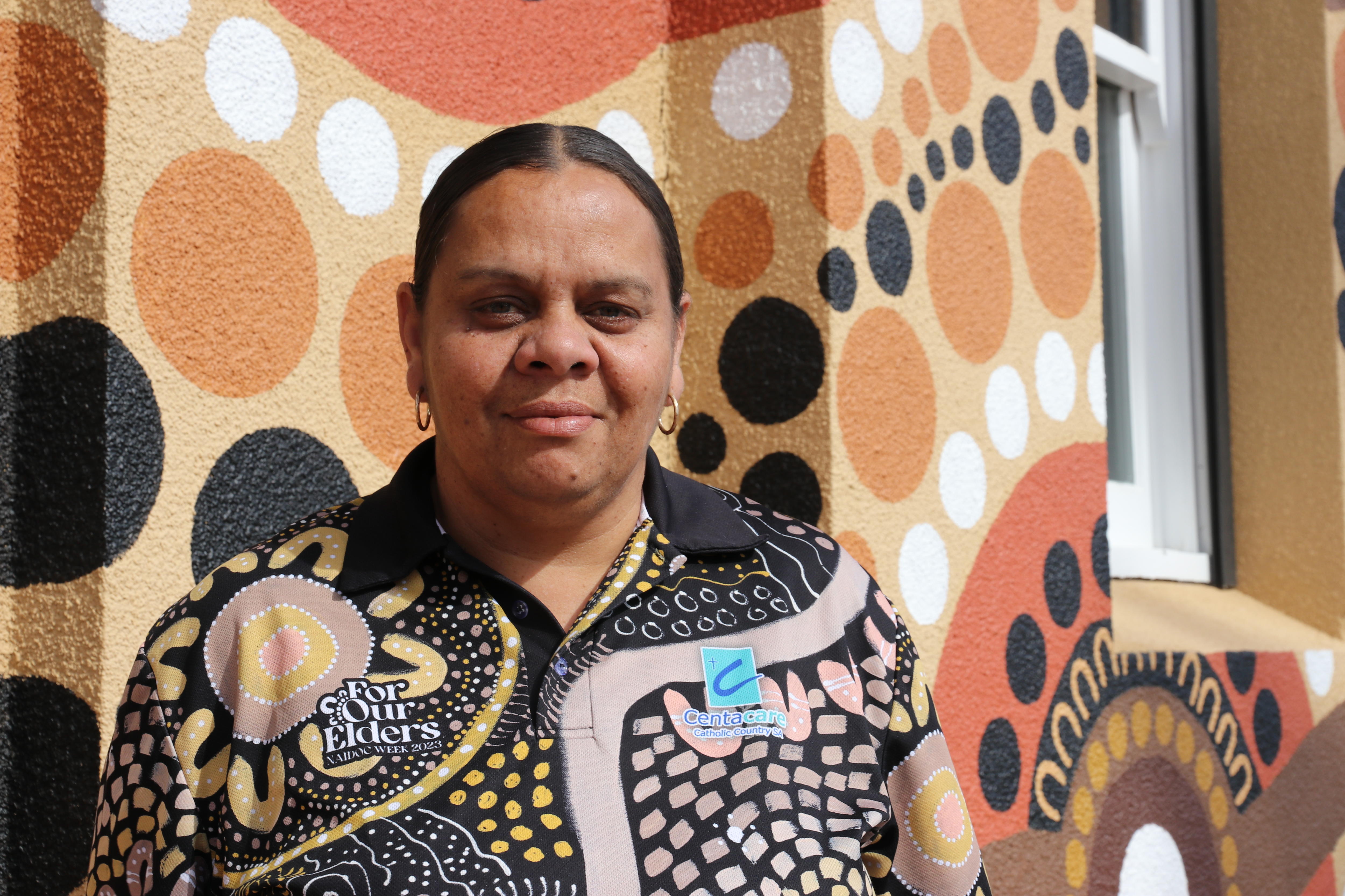 Woman stands in front of Indigenous dot painting mural on building smiling to camera