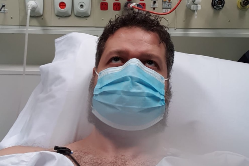 Man wearing a blue mask and white gown lies in hospital bed with equipment behind him