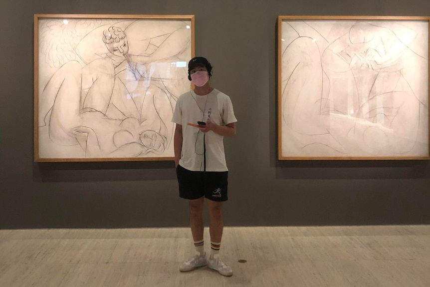 Will standing in an art gallery wearing a mask and holding his phone with earphones in.