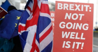 Protest signs that read 'Brexit: Not going very well, is it!' stand beside a road with British flags