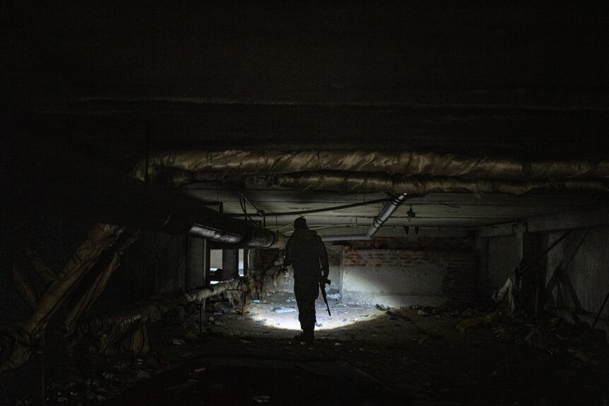 A soldier holding a rifle shines a torch as he walks through an underground basement.