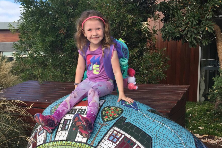 girl sits on mosaic rock in front of kindergarten smiling