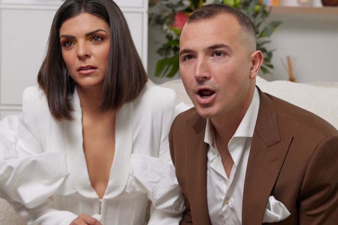 Steph and Gian sit on a couch looking shocked as their auction takes place.