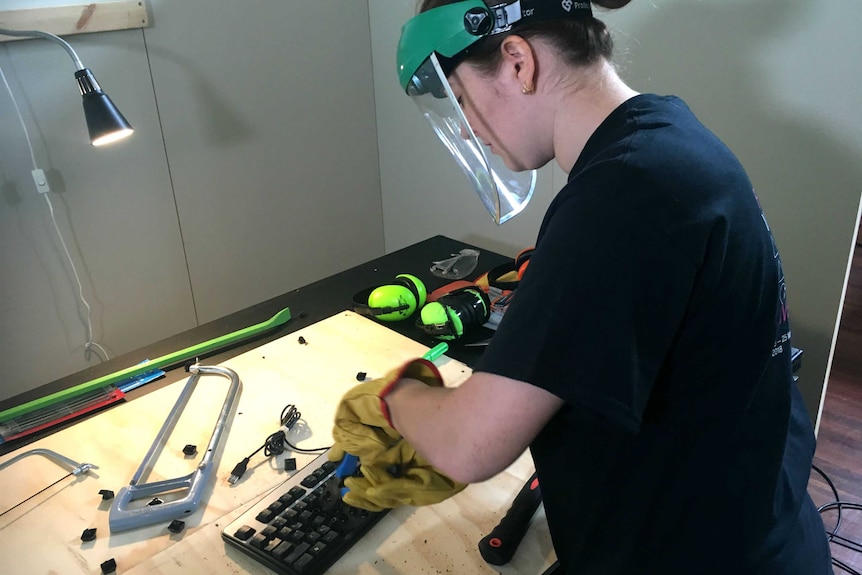 A woman wearing safety equipment uses pliers to pull apart a computer keyboard.