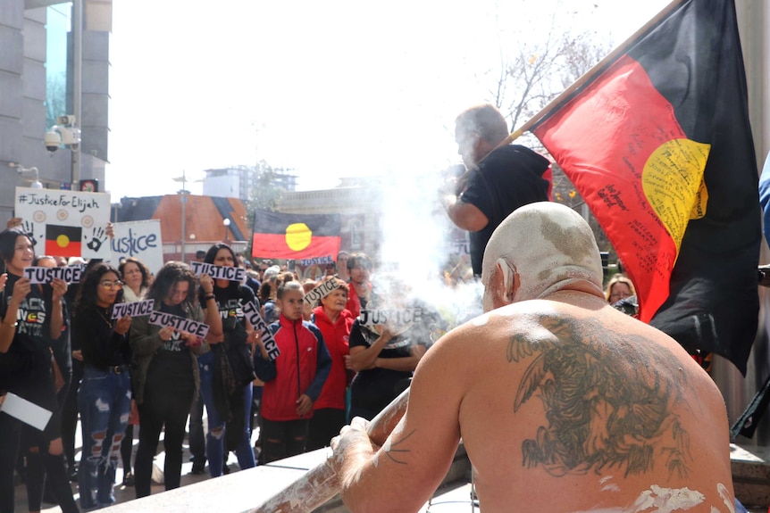 A man carrying an Aboriginal flag addresses a large crowd, with people holding Justice for Elijah signs.