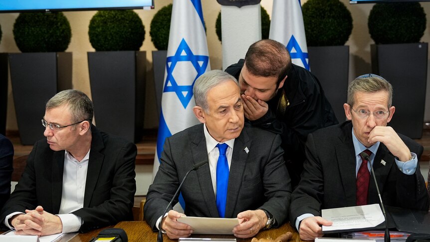 Benjamin Netanyahu sits at a table with two men sitting either side of him as another man whispers in his ear