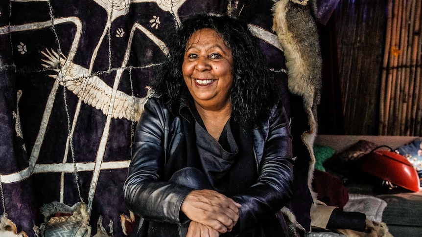 A 60-something Aboriginal woman sits smiling in her studio with an embroidered tapestry behind her
