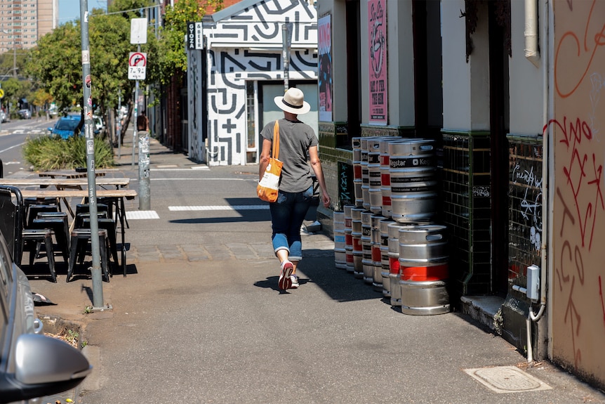 Mujer con sombrero y bolso camina por la calle pasando por un pub y una hilera de barriles de cerveza plateados