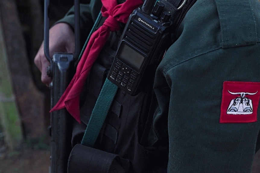 A close up of a person shows a red and white badge on their green jacket, their gun and their radio device