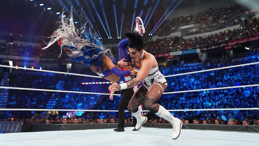 A woman with black hair holds another woman mid-air during the middle of slamming her to the ground.