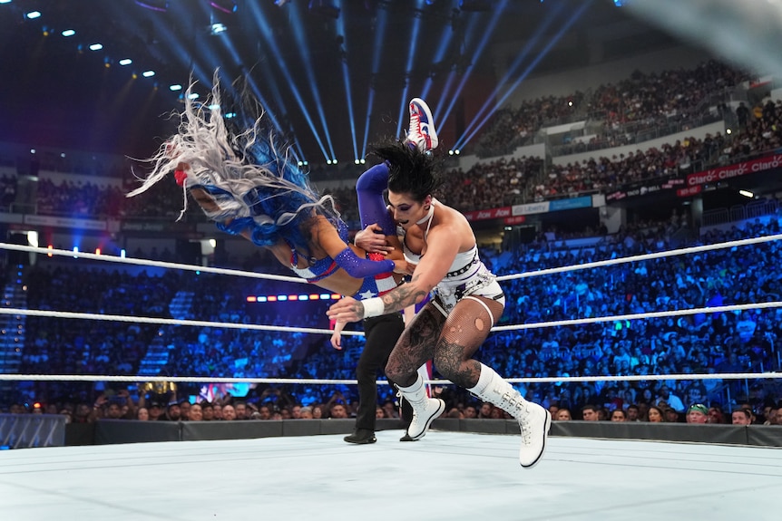A woman with black hair holds another woman mid-air during the middle of slamming her to the ground in a ring.