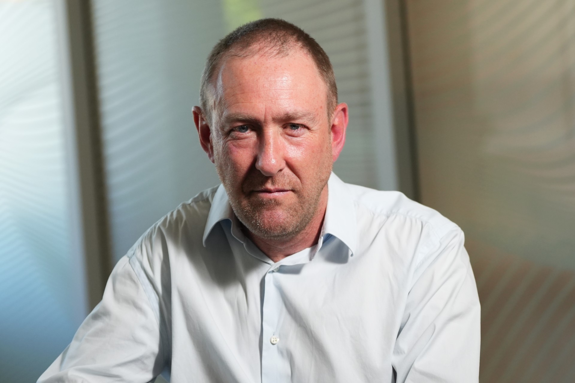 A man in a light blue longsleeve shirt looks with a neutral expression at the camera while indoors.