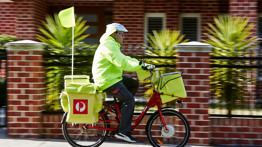 Un cartero viste ropa de alta visibilidad mientras entrega el correo en una bicicleta eléctrica, con una casa de ladrillos en el fondo.