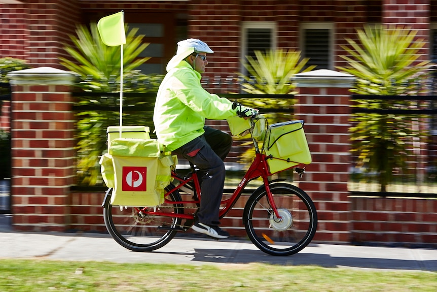 A postman wears high-vis clothing while delivering mail on an electric bike, brick house in background.