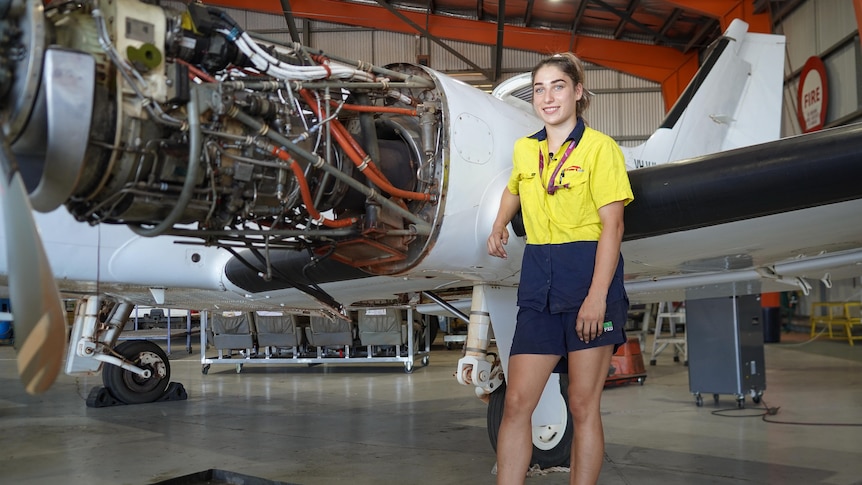 A woman in high-vis workwear stands next to a small airplane with panels stripped back to show the aircraft's wiring.