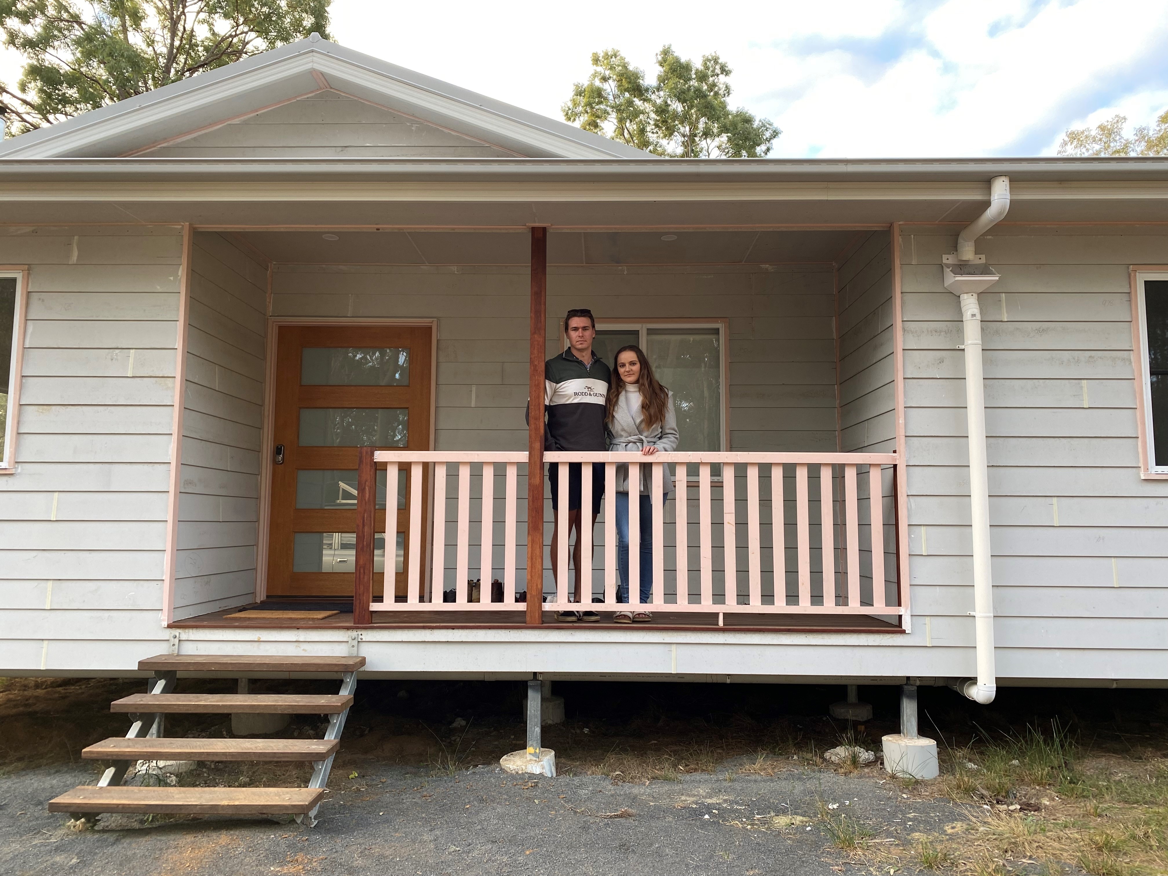 A young couple stand on the verandah of their weatherboard home.