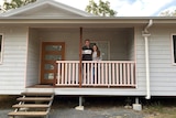 A young couple stand on the verandah of their weatherboard home.