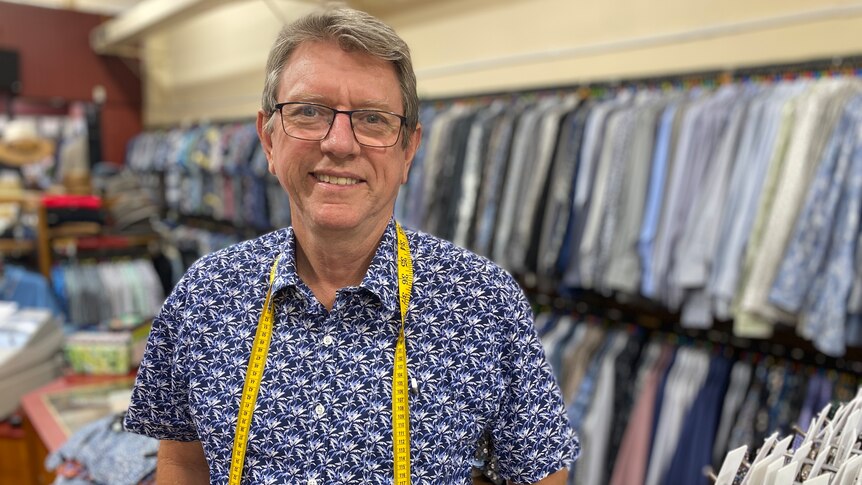 man stands with a measuring tape around his neck with racks of clothing behind him