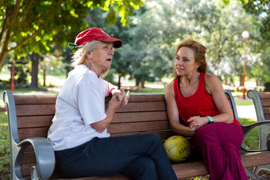 woman wearing red singlet and long pants chats with another woman wearing a hat sitting on a park bench