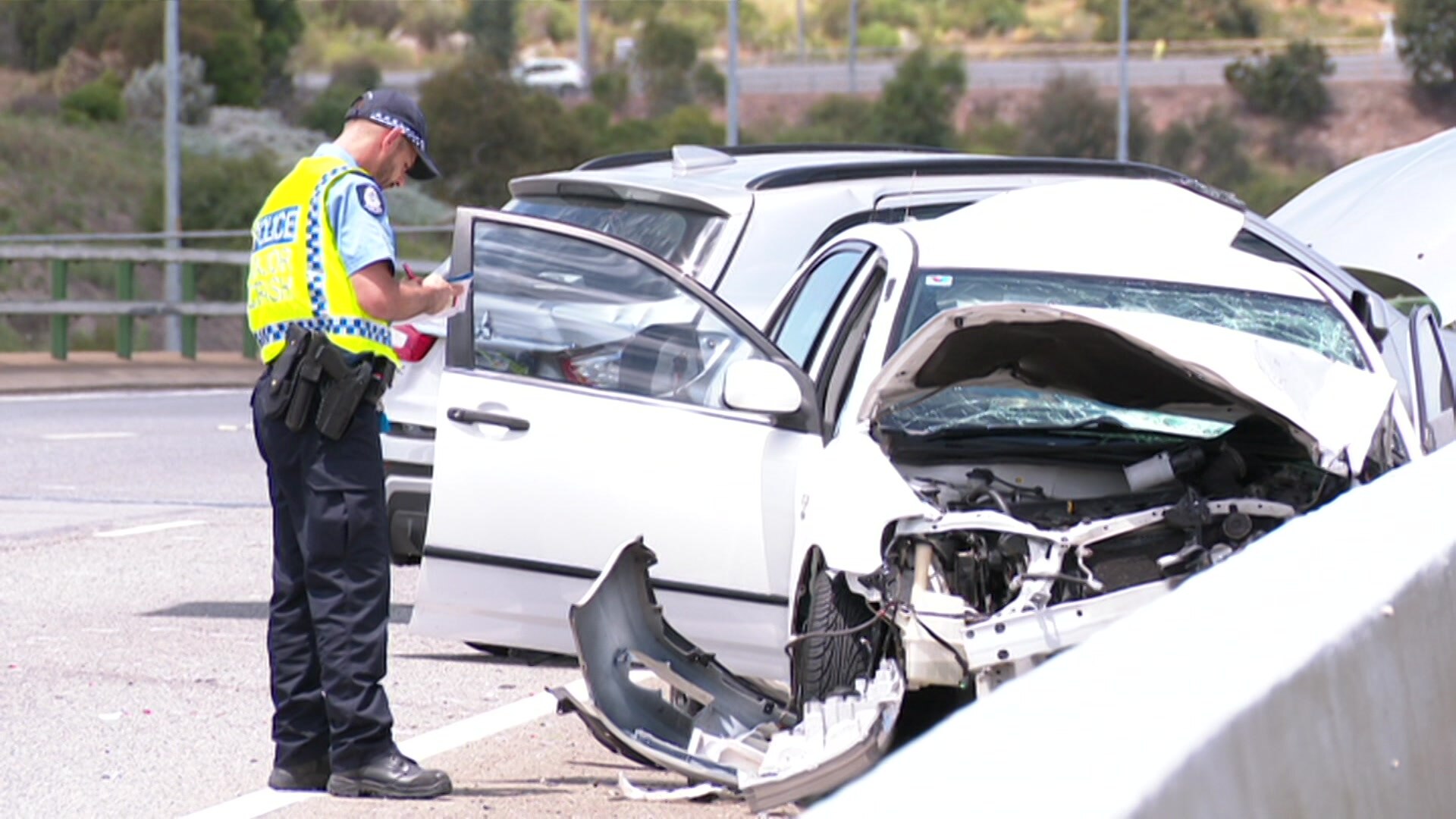 A police officer stands next to a white car that is smashed up at the front
