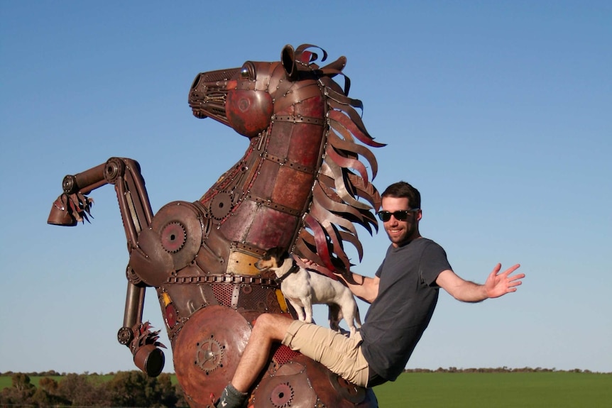 A man in sunglasses grinning as he sits astride a sculpture of a large metal horse.