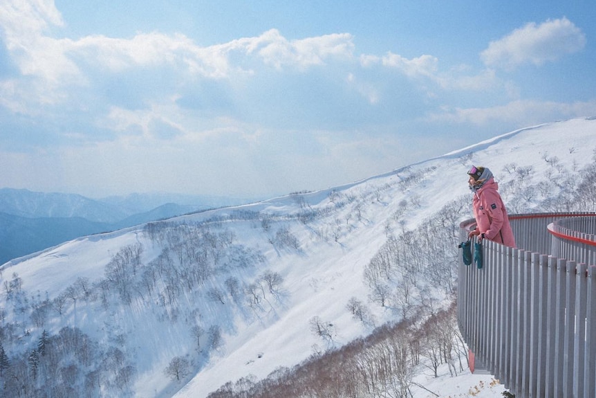 Rani is seen standing on a platform and looking out to a mountain on her left while wearing snowboarding gear on a sunny day.
