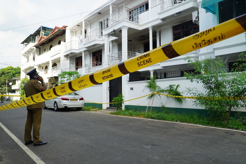 Yellow crime scene tape blocks off a white house as a police man stands and talks on his phone.