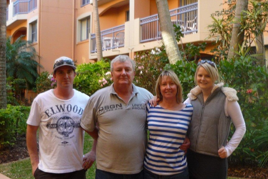A family of four- parents and two adult children- stand in front of a set of units lines with trees