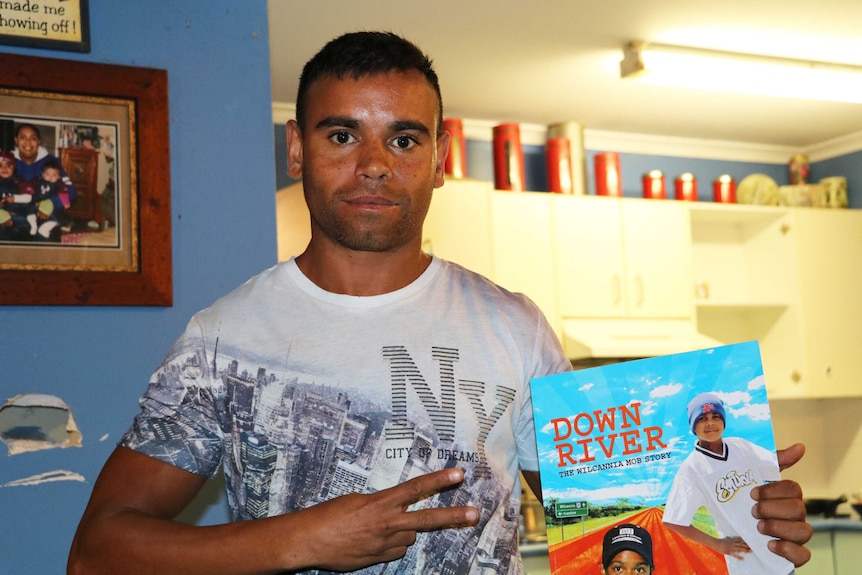 Photo of Buddy Blair standing in a kitchen, holding up a book about the Wilcannia Mob.