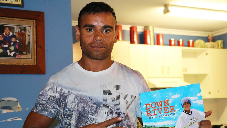 Photo of Buddy Blair standing in a kitchen, holding up a book about The Wilcannia Mob.