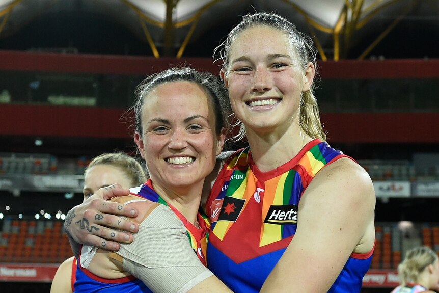 Daisy Pearce and Tayla Harris of the Melbourne Demons smile for the camera while hugging after an AFLW game.