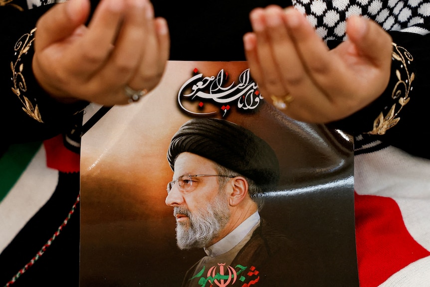 Two hands pray in front of the picture of the late Iranian President Ebrahim Raisi, who is wearing a turban and glasses.