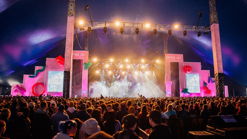 A festival crowd under a large tent in front of a pink Spin Off stage where The Chats are playing under lights.