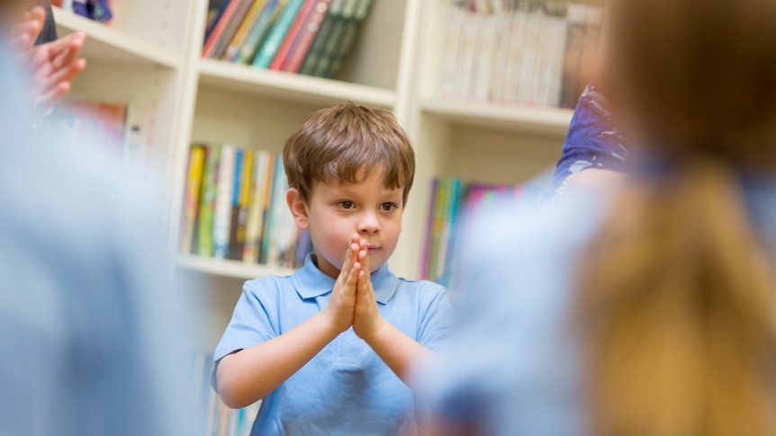 young boy in school uniform with hands together with bookshelves in background