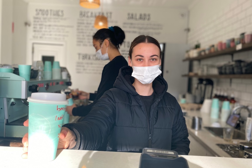 Woman in black jacket wearing face mask, holding out a green takeaway coffee cup
