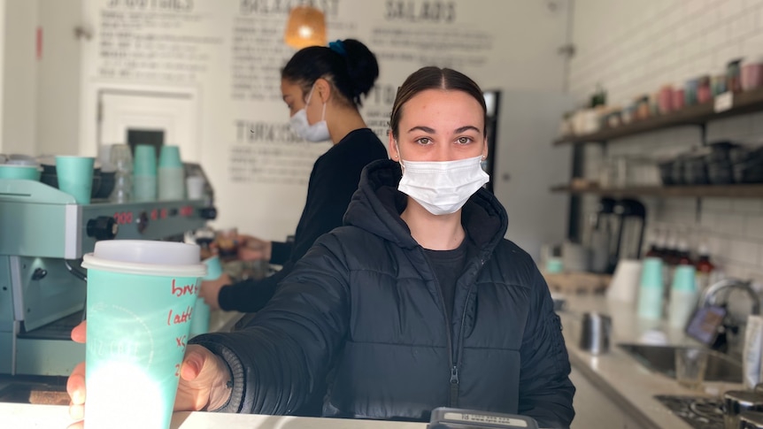 Woman in black jacket wearing face mask, holding out a green takeaway coffee cup