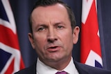 Mark McGowan in a blue suit, standing in front of a blue background and Australian flags.