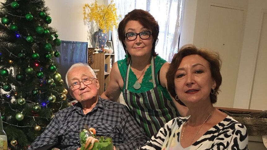 An older man sits at a Christmas lunch table with two women and a decorated tree behind them